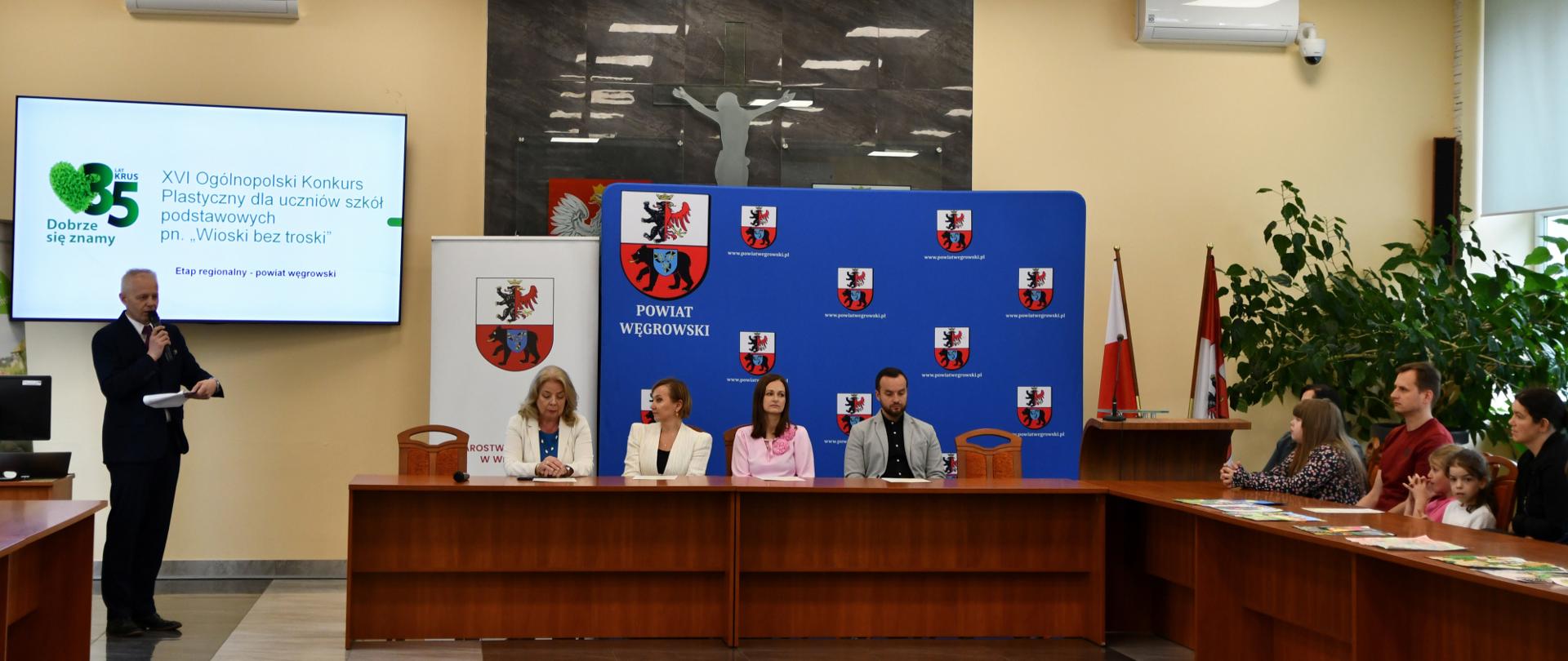W sali konferencyjnej stoi mężczyzna z mikrofonem przed ekranem telewizora, na prezydium zasiadają trzy kobiety i mężczyzna