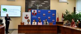W sali konferencyjnej stoi mężczyzna z mikrofonem przed ekranem telewizora, na prezydium zasiadają trzy kobiety i mężczyzna