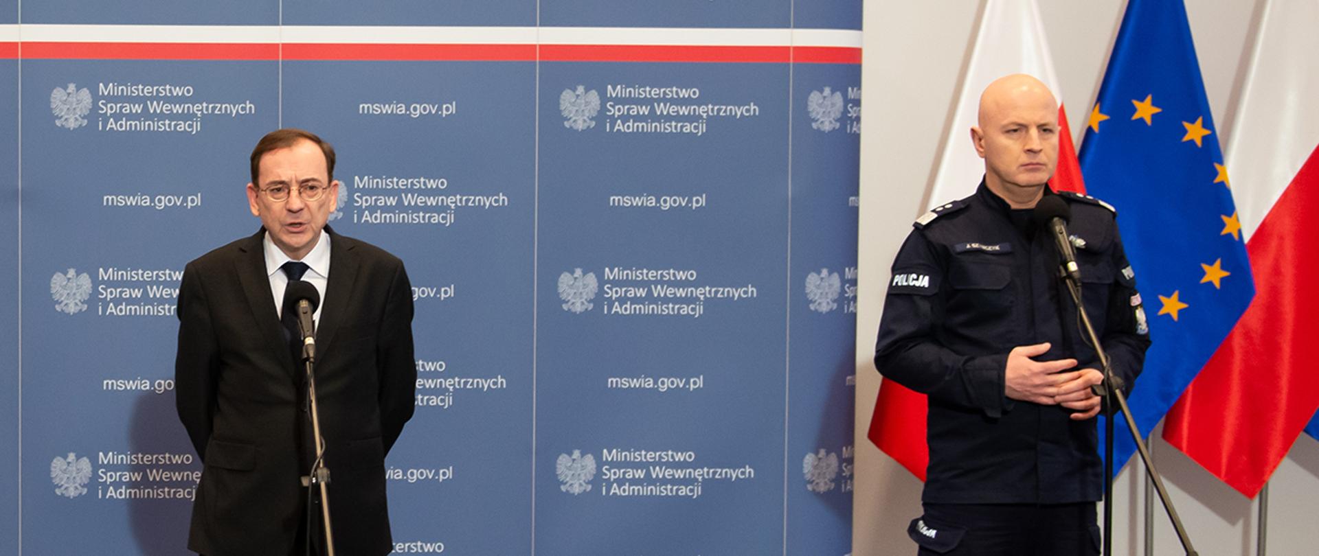Na zdjęciu widać stojących przy mikrofonach Mariusza Kamińskiego ministra SWiA i gen. insp Jarosława Szymczyka. Minister przemawia. W tle widać fragment ścianki MSWiA i flagi narodowe Polski i UE.