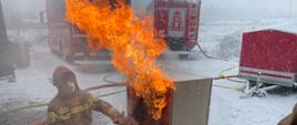 Strażak przedstawia symulacje pożar domu na podstawie makiety widoczne płomienie w tle dwa samochody pożarnicze oraz czerwona przyczepka strażak klęczy przy makiecie ma maseczkę na twarzy obok niego worek