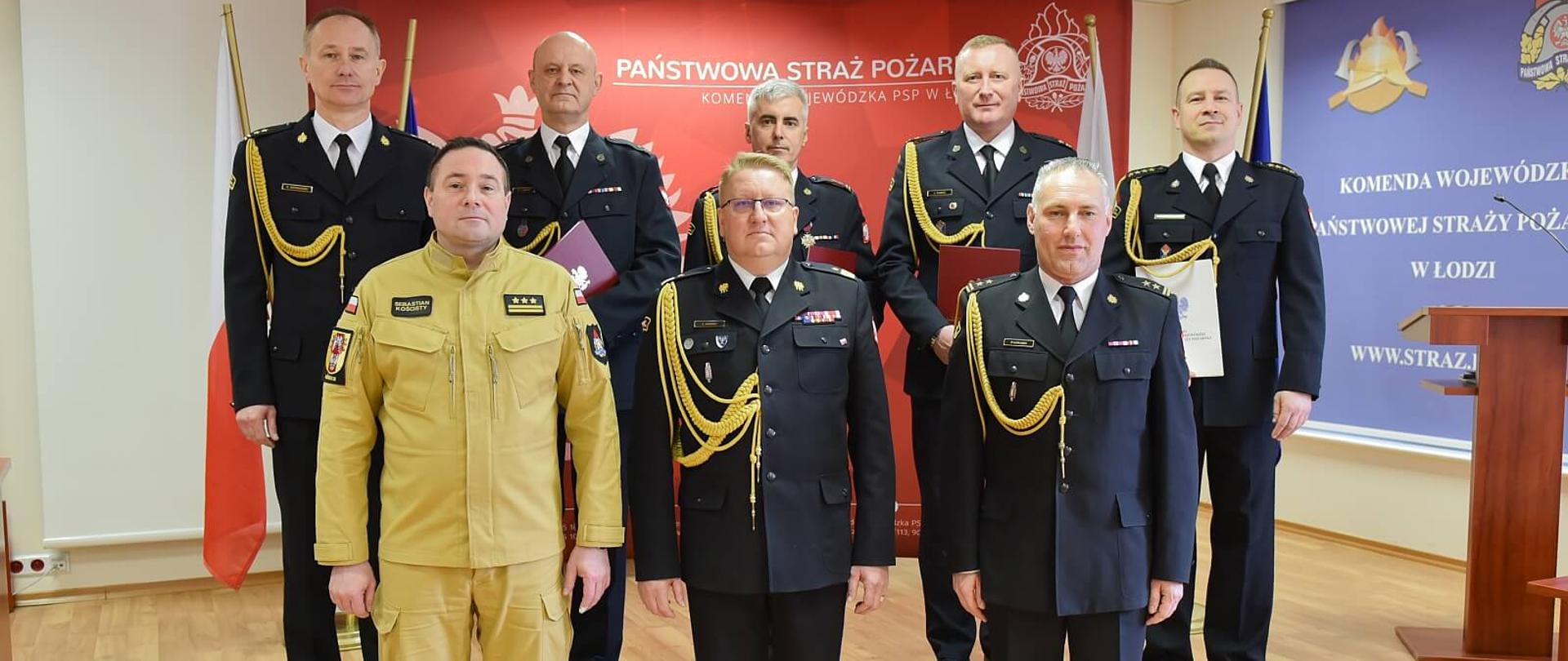 na zdjęciu 8 strażaków pozuje do zdjęcia na tle baneru z napisem Państwowa Straż Pożarna Komenda wojewódzka w Łodzi, jeden ze strażaków ma mundur w kolorze pisakowym reszta jest w mundurach wyjściowych