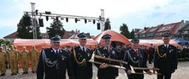 Zdjęcie przedstawiające pięciu strażaków stojących w rzędzie na wybrukowanym placu. Funkcjonariusze są ubrani w ciemne mundury galowe z licznymi odznaczeniami, baretkami i sznurami galowymi. Dwaj strażacy w środku trzymają ozdobne, pamiątkowe topory strażackie umieszczone na drewnianych podstawach. W tle widoczny jest duży pomarańczowy namiot, metalowa konstrukcja sceniczna z oświetleniem oraz rzędy innych strażaków, w tym grupa w piaskowych mundurach służbowych. Nad placem górują zabytkowe kamienice.