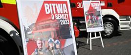 Dwa plakaty promocyjne akcji "Bitwa o Remizy 2023"; na tle dwóch pojazdów pożarniczych.