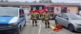 Strażacy czekają na parkingu przed budynkiem ze sprzętem do udzielania pierwszej pomocy do budynku. Za nimi stoi samochód pożarniczy.