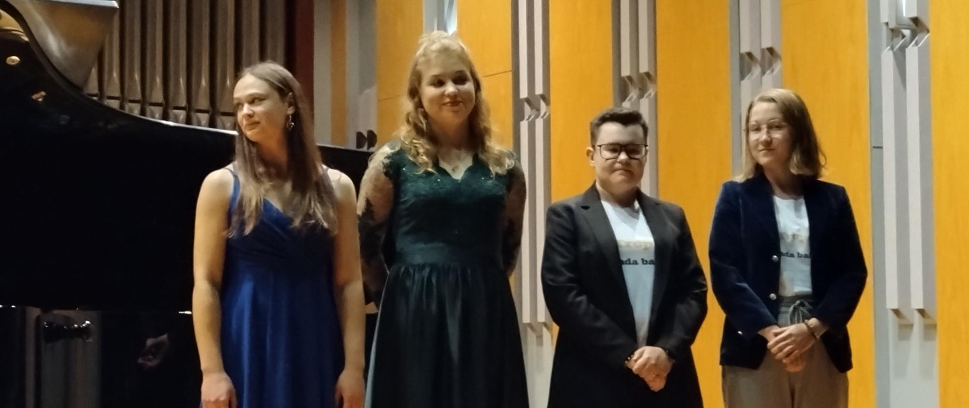 Grupowe zdjęcie. Wykonawcy stoją w jednym rzędzie na scenie sali koncertowej, za nimi fortepian z podniesiona klapą. W tle organy oraz jedna ściana sali koncertowej w szaro-pomarańczowym kolorze.