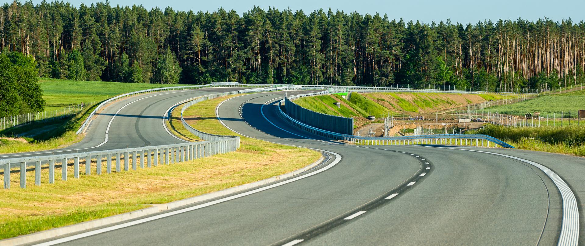 Dwie równoległe jezdnie drogi ekspresowej S61 w zakrętach w lewo i w głębi w prawo. Na drodze pusto. 