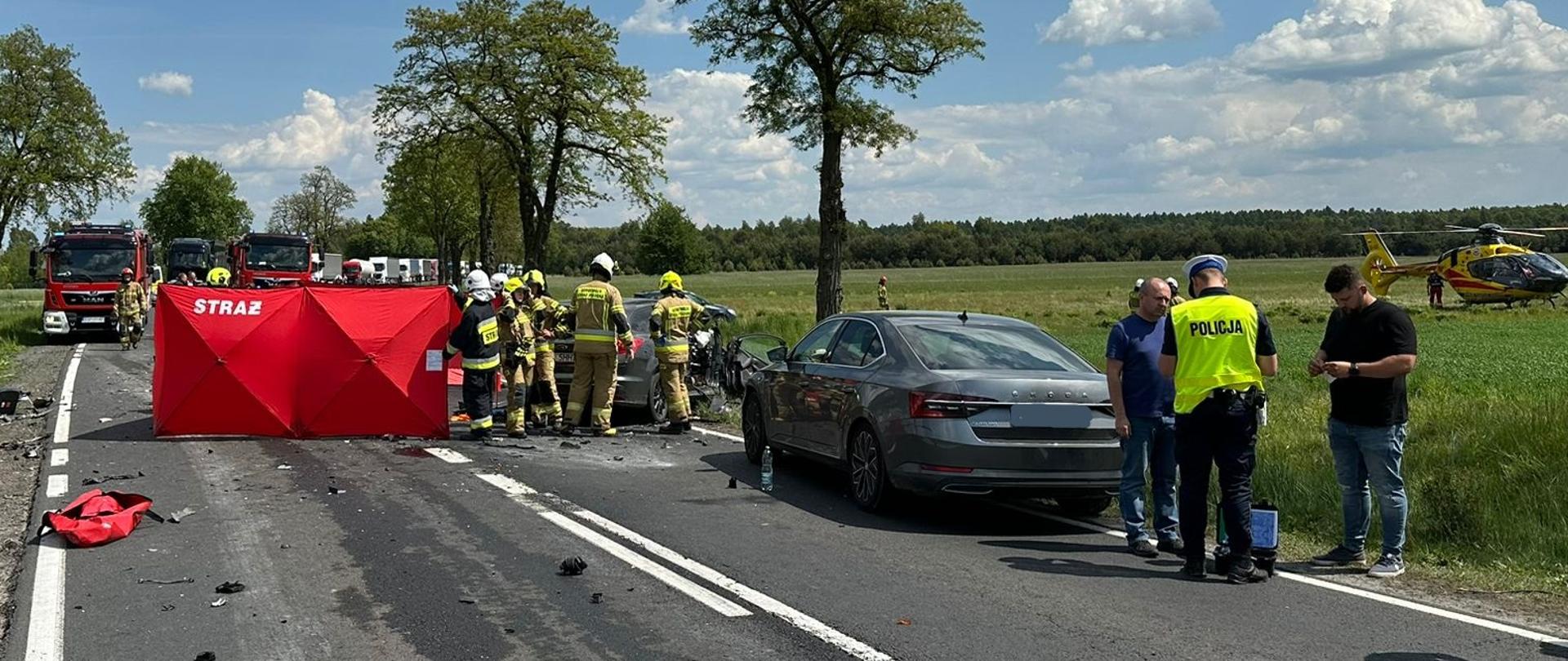 Wypadek drogowy w miejscowości Strzelce. Na drodze stoją uszkodzone auta uczestniczące w wypadku. W akcji biorą udział policjanci oraz straż pożarna. Na środku drogi czerwony parawan zasłaniający poszkodowane osoby. W tle wozy strażackie. Z prawej strony śmigłowiec LPR.