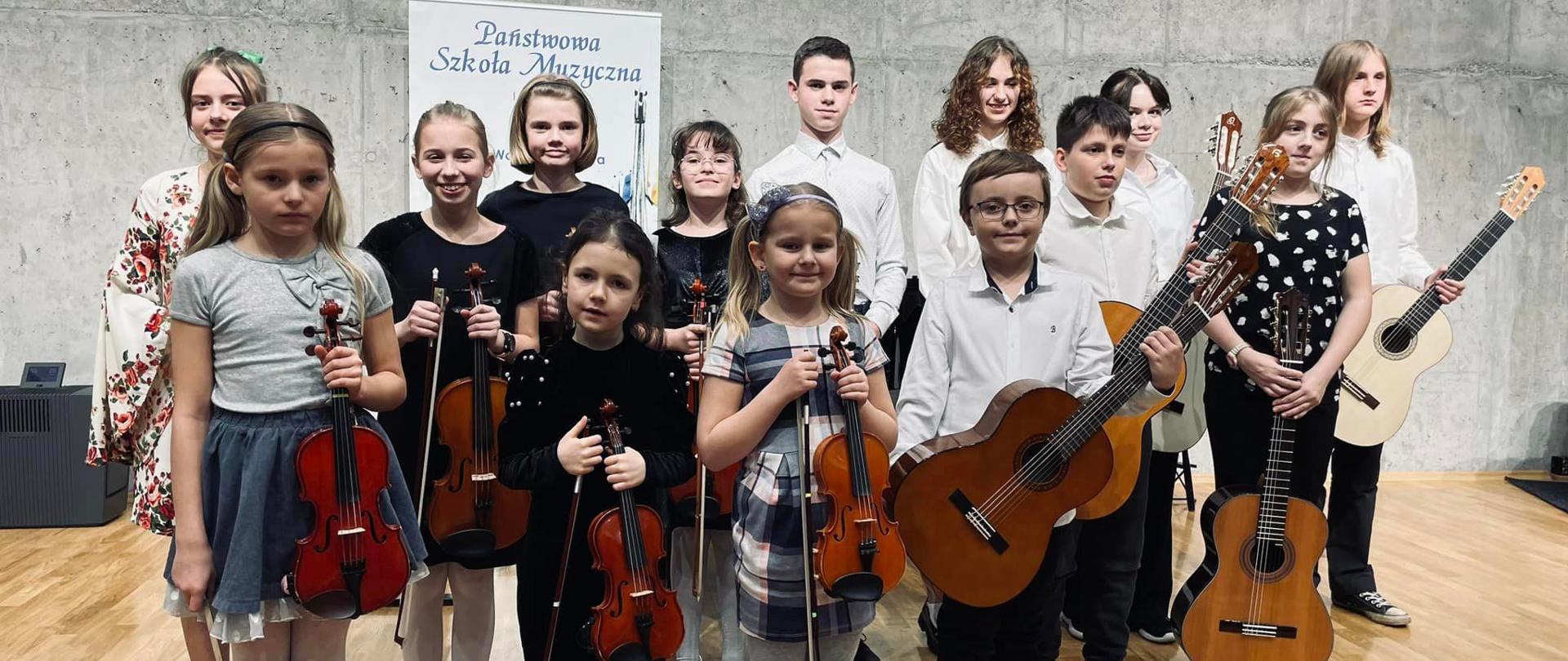 Grupa dzieci na scenie sali koncertowej trzyma w rękach skrzypce i gitary