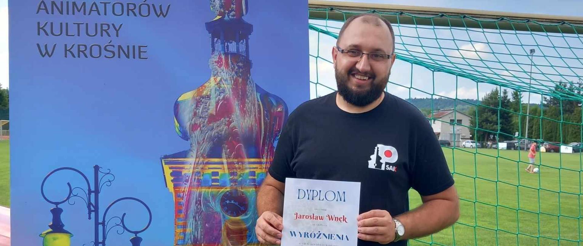 Słuchacz Studium stoi na tle banneru reklamowego studium i trzyma dyplom z wyróżnieniem. 