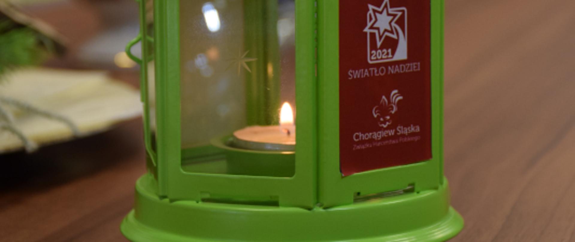 Zielona latarenka, w środku widoczny płomyk Betlejemskiego Światełka Pokoju, na latarence na czerwonym tle napis "Betlejemskie Światełko Pokoju Światło Nadziei, Chorągiew Śląska 