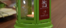 Zielona latarenka, w środku widoczny płomyk Betlejemskiego Światełka Pokoju, na latarence na czerwonym tle napis "Betlejemskie Światełko Pokoju Światło Nadziei, Chorągiew Śląska 
