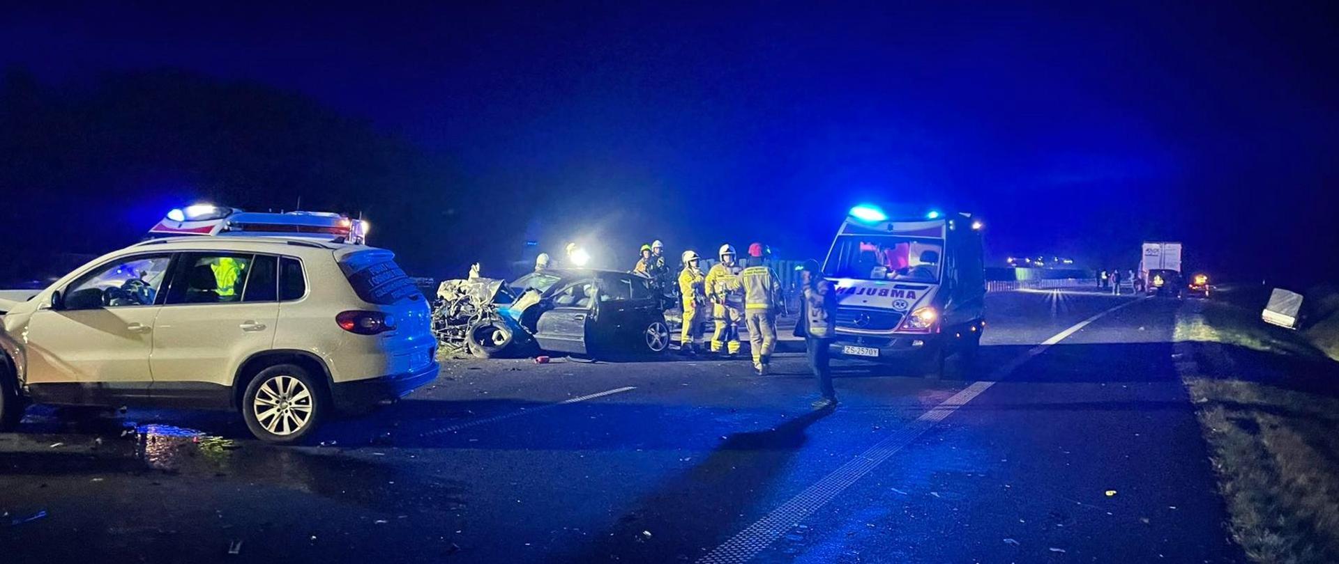 Na zdjęciu widoczne uszkodzone 2 samochody osobowe koloru czarnego i białego. Po bokach pojazdów stoją 2 białe pojazdy Państwowego Ratownictwa Medycznego, mają włączoną sygnalizację ostrzegawczą koloru niebieskiego. Obok czarnego samochodu stoją strażacy w ubraniach specjalnych koloru piaskowego. W tle inne rozbite pojazdy uczestniczące w zdarzeniu. Pora nocna.