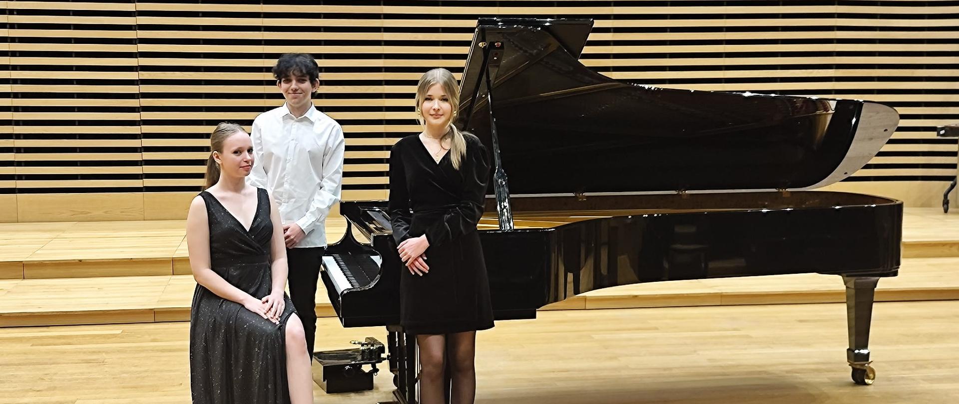 Dwie dziewczyny i chłopak stoją przy fortepianie na scenie.