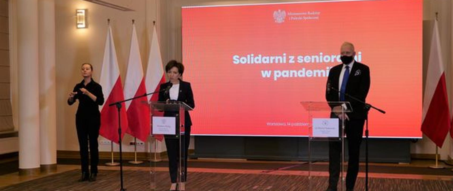 Sala. Minister Maląg stoi przy mównicy. Uczestniczy w brefingu. Z tycłu baner z napisem "Solidarni z seniorami w pandemii".