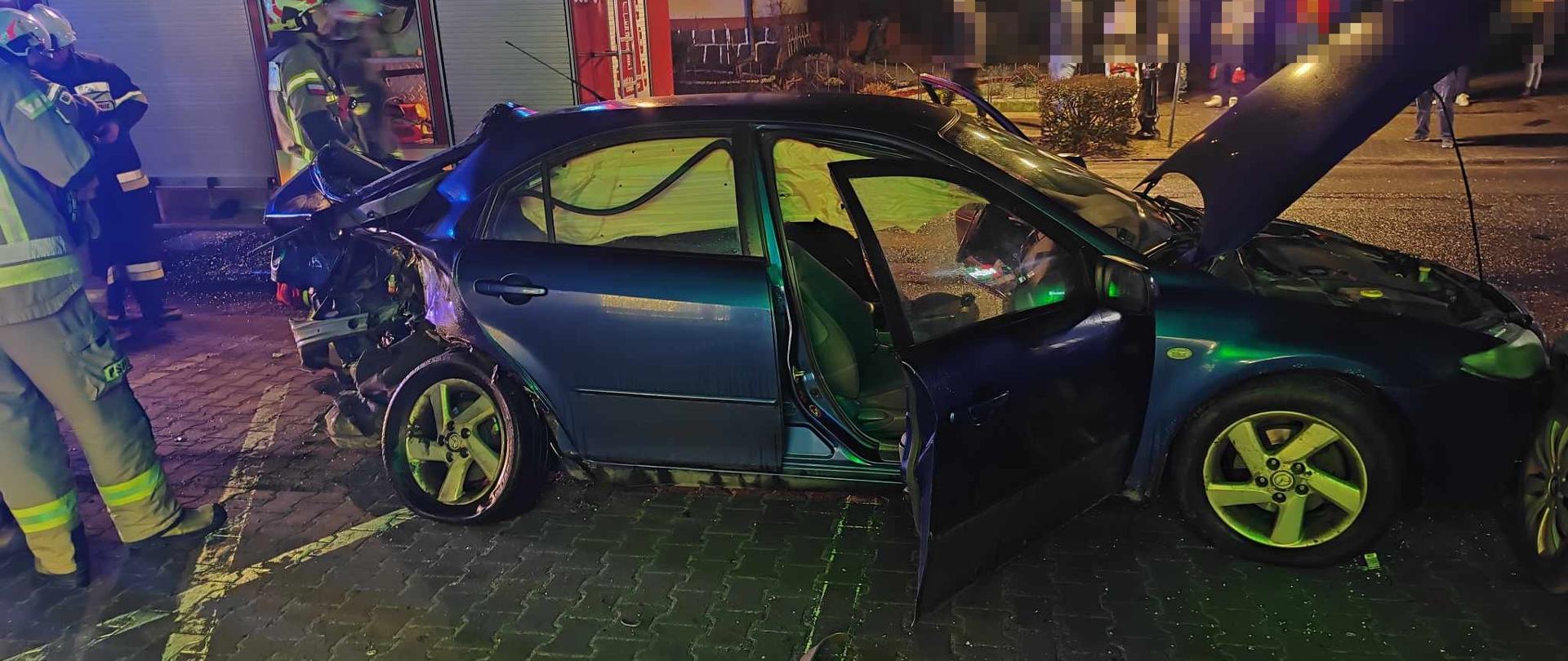 Na zdjęciu widać uszkodzony pojazd osobowy w tle wóz strażacki i strażaków 