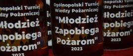 Eliminacje gminne OTWP w Jutrosinie. Sala w siedzibie Ochotniczej Straży Pożarnej w Jutrosinie. Na stole ustawione są kubki - nagrody dla uczestników, na których widnieje napis "Ogólnopolski Turniej Wiedzy Pożarniczej "Młodzież Zapobiega Pożarom" 2023.