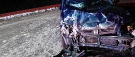 Na pierwszym planie widać rozbity przód samochodu osobowego. Auto stoi na ośnieżonej jezdni w porze nocnej. Na drugim planie widać posesję prywatną.