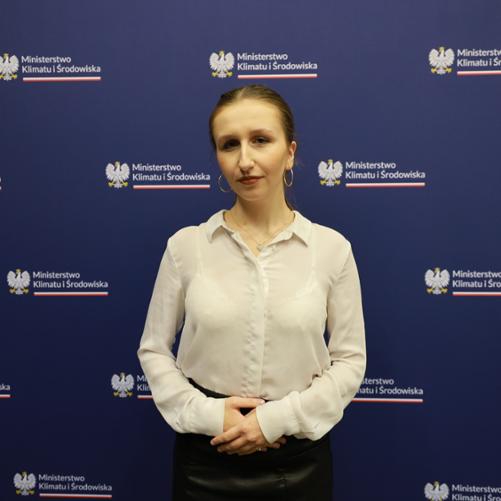 Na zdjęciu znajduje się młoda kobieta na niebieskim tle z logo Ministerstwa Klimatu i Środowiska. Na uszach znajdują się złote kolczyki w kształcie kół. Włosy w kolorze ciemnego blondu uczesane są w kucyk.