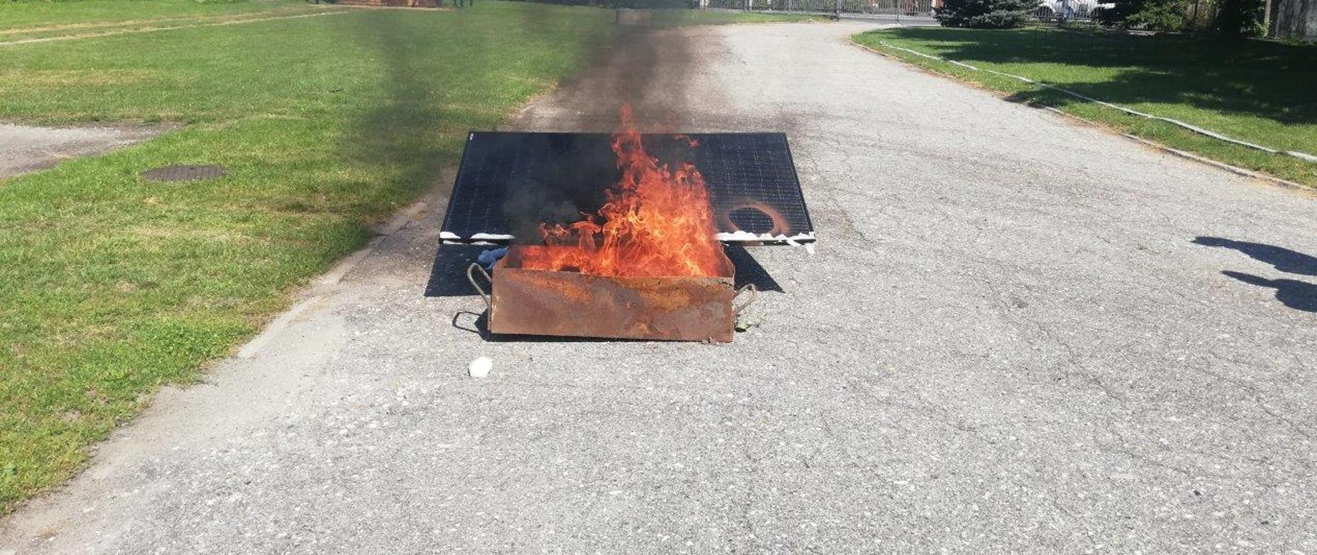 Na placu komendy powiatowej celem demonstracji pożaru paneli fotowoltaicznych podpalono panel fotowoltaiczny. Do tego celu utworzono ognisko w metalowej kuwecie. Panel topi się pod wpływem temperatury. Nad panelem unosi się czarny dym. 