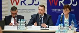 Za stołem siedzi minister Czarnek, kobieta w niebieskiej marynarce i mężczyzna w garniturze, minister mówi do stojącego przed nim mikrofonu, za nimi banery z napisem WRDS.
