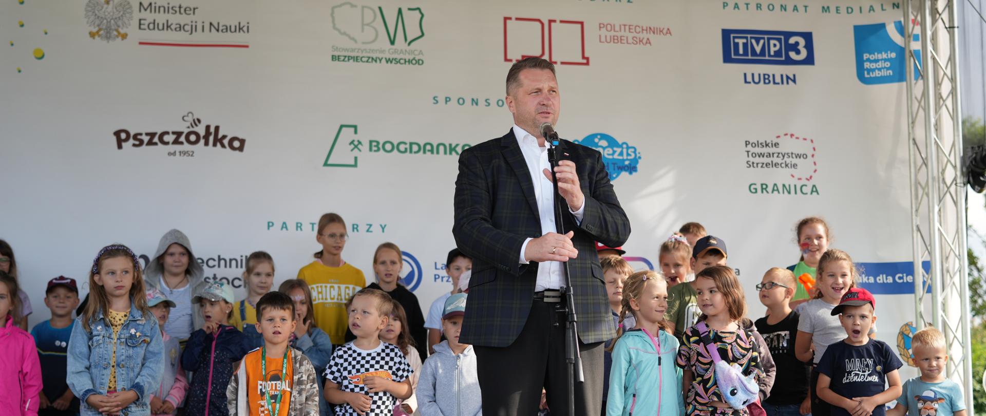 Minister Czarnek stoi i mówi do mikrofonu, za nim na estradzie z stoi duża grupa kolorowo ubranych dzieci.