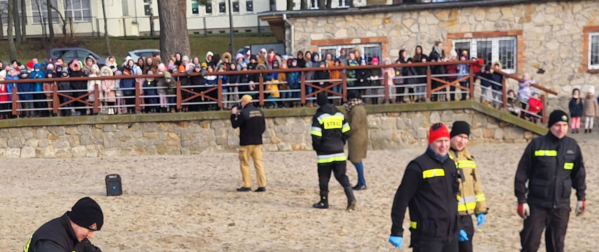 pora dzienna, widać plażę- na niej funkcjonariuszy PSP. Na promenadzie widać dużo uczniów.