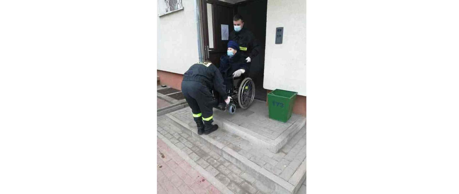 Dwóch strażaków znoszących kobietę na wózku inwalidzkim z klatki bloku mieszkalnego.