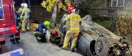 Zdjęcie przedstawia rozbity samochód osobowy oraz strażaków zabezpieczających miejsce zdarzenia.