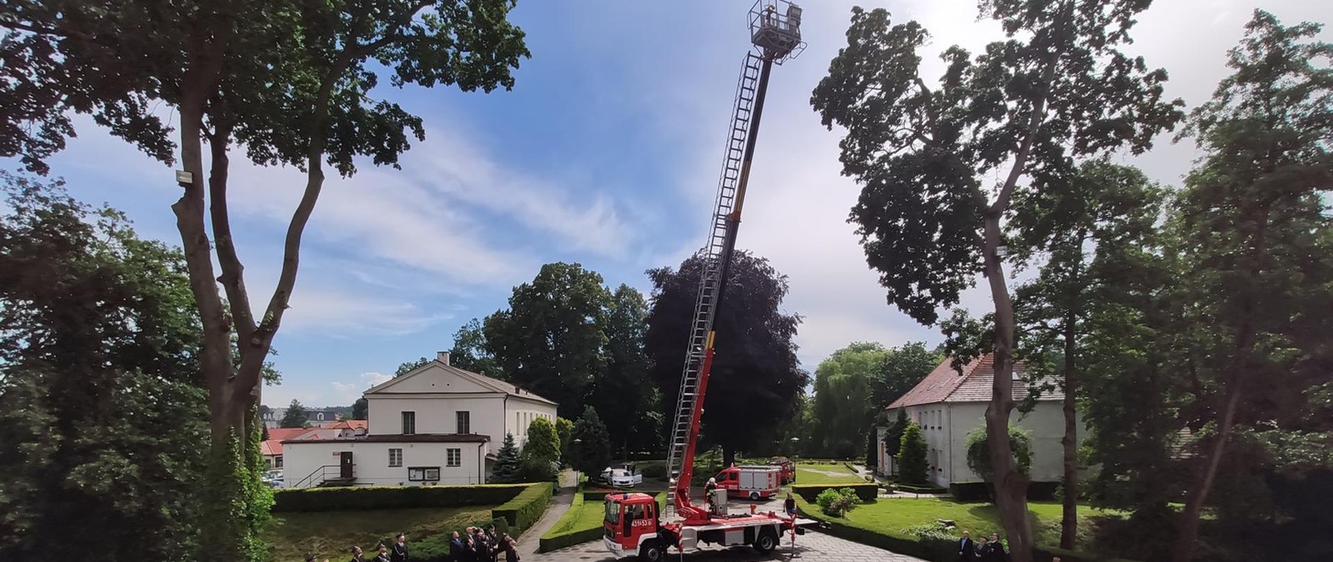 Na zdjęciu widoczny strażacki pojazd bojowy z wysuniętym podnośnikiem z koszem oraz osoby oglądające.
