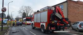 Na środku jezdni stoi uszkodzony samochód osobowy oraz autobus MZK. Przy samochodzie osobowym stoją strażacy. Na zdjęciu widać również samochód strażacki, radiowóz oraz karetki pogotowia ratunkowego. 