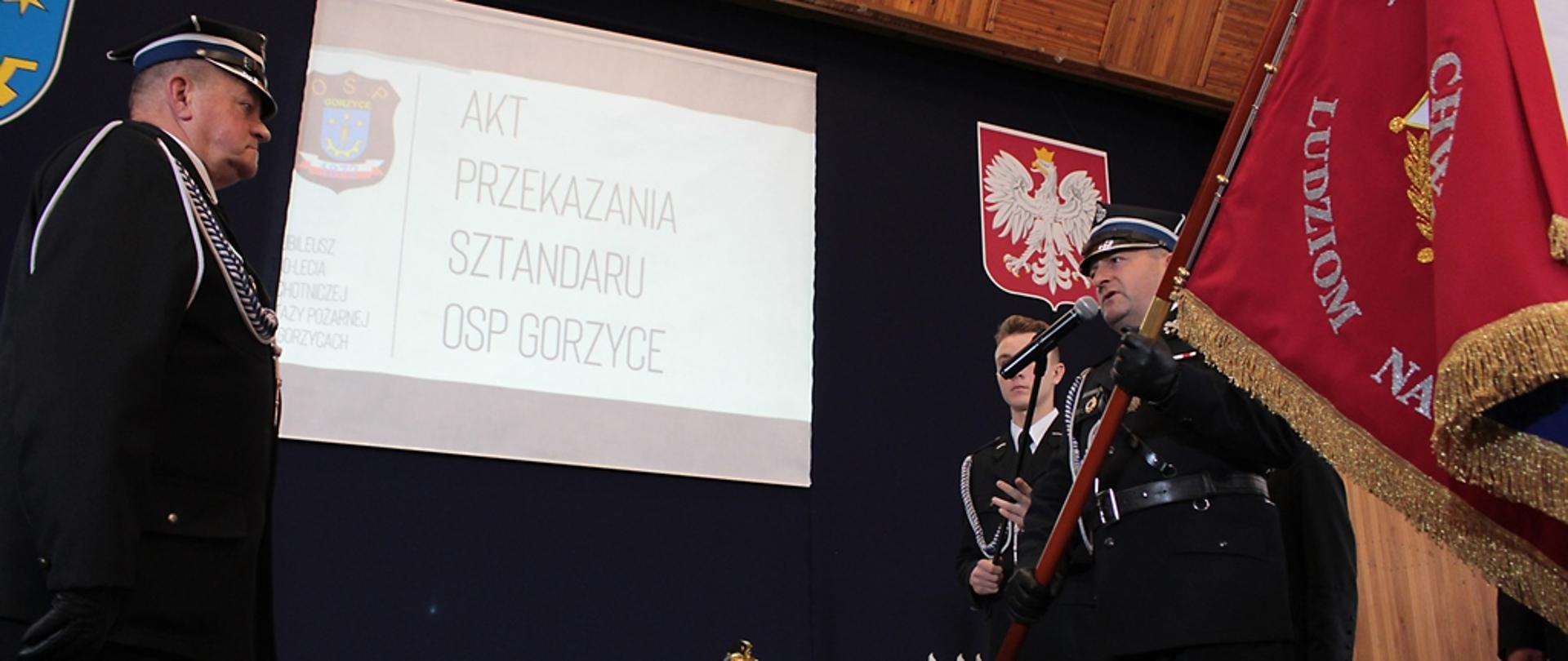 Strażak stoi przed mikrofonem i trzyma sztandar z zamiarem przekazania go innemu strażakowi, który stoi naprzeciw niego. 