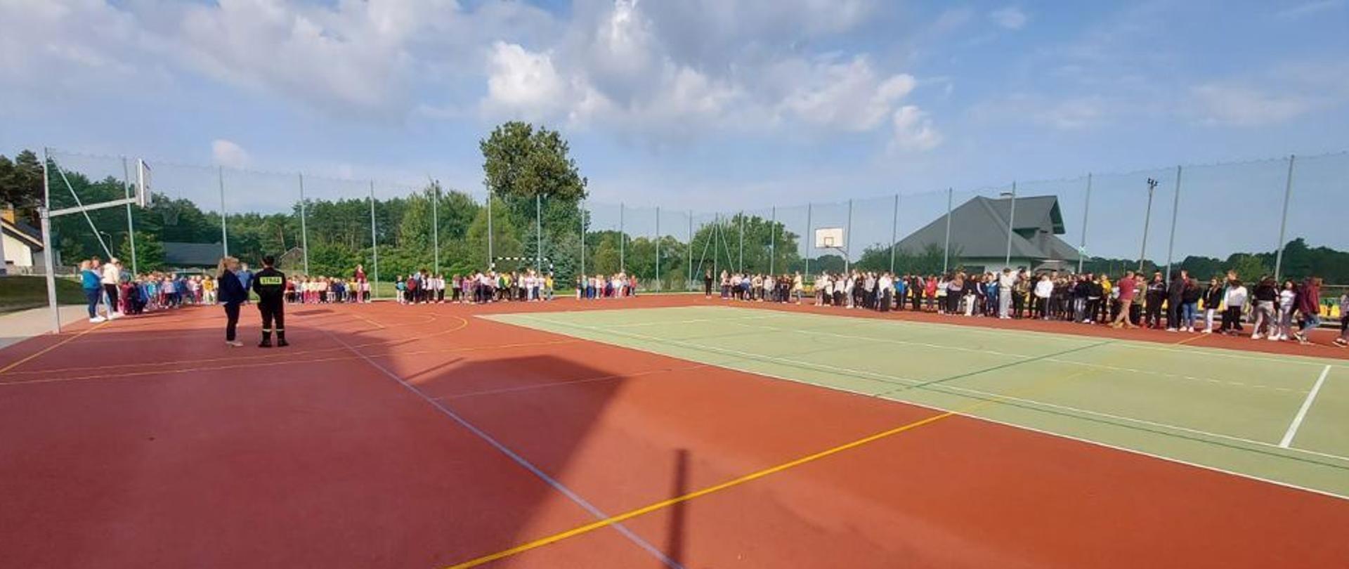 Boisko szkolne, dyrektor placówki zdaje relację z przeprowadzonej ewakuacji funkcjonariuszowi, na drugim planmie zebrani uczniowie w miejscach wyznaczonych