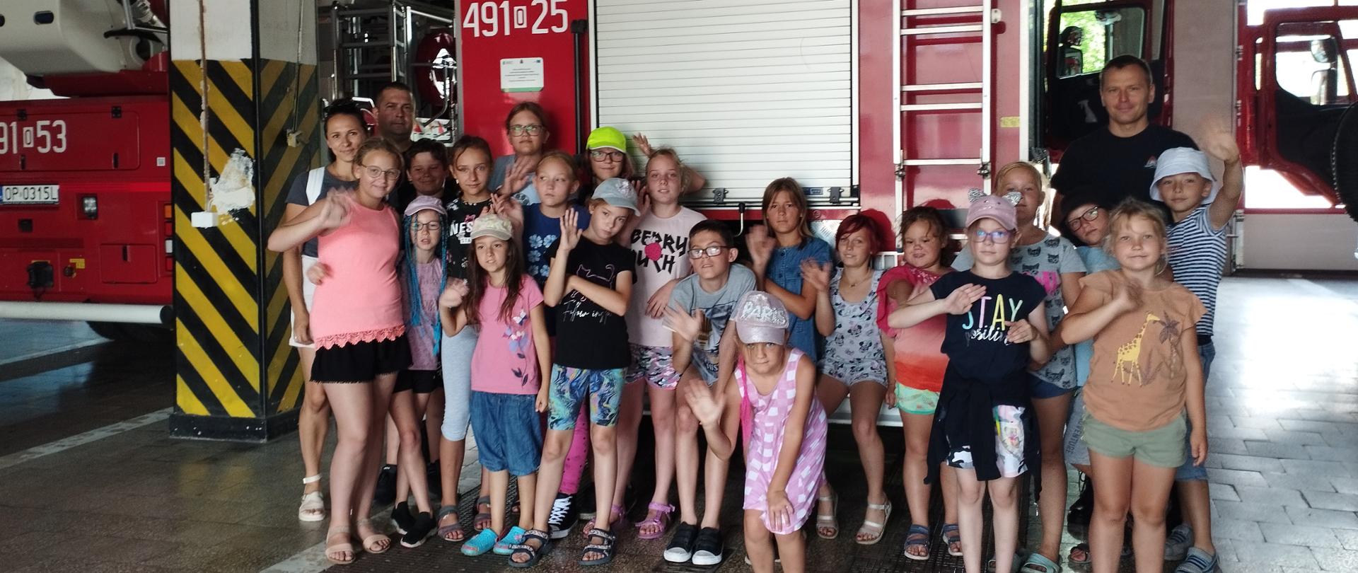 Na zdjęciu znajdują się dzieci oraz strażak i opiekun ustawieni na garażu na tle samochodu strażackiego