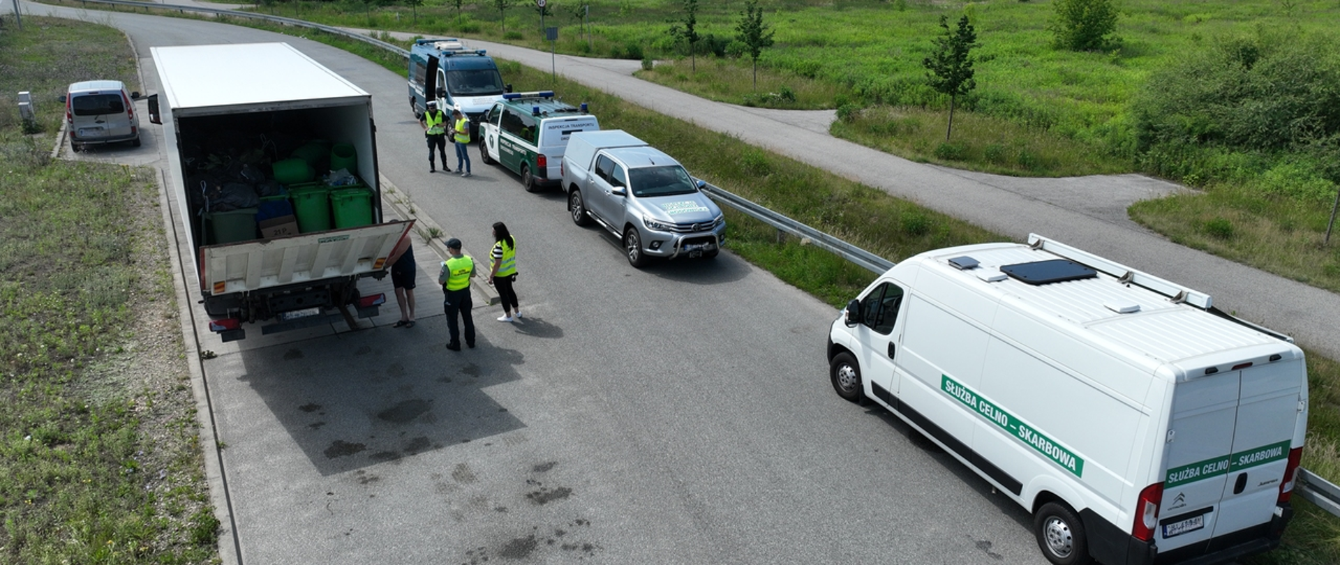 Inspektorzy WIOŚ w Warszawie kontrolują ciężarówkę. Kierowca otwiera drzwi naczepy, wewnątrz widać liczne pojemniki. Na poboczu stoją samochody współpracujacych służb.