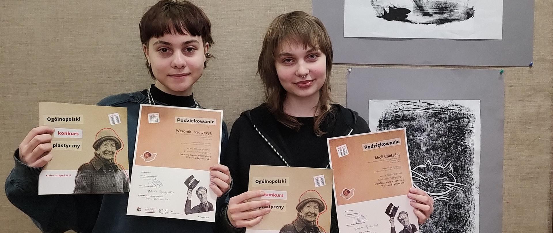 Na zdjęciu dwie uczennice prezentujące podziękowania za udział w konkursie i katalogi wystawy pokonkursowej, w tle prace konkursowe uczennic