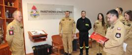 Komendant Powiatowy PSP w Golubiu-Dobrzyniu trzyma w dłoniach teczkę w kolorze czerwonym, przed nim stoi st. ogn. Radosław Dulczewski.