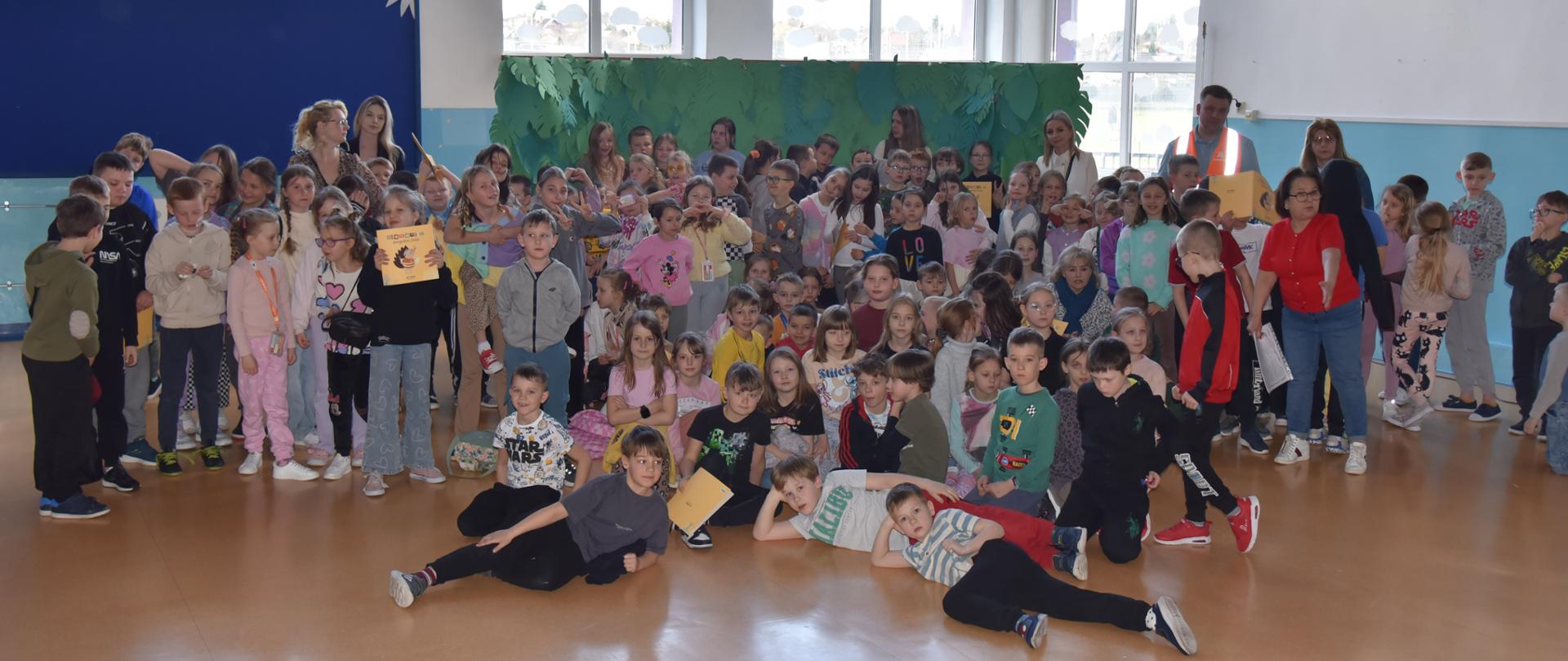 Grupa uczniów Szkoły Podstawowej nr 9 w Ełku pozuje wspólnie do zdjęcia w holu. Dzieci w różnym wieku stoją i siedzą razem,, niektóre trzymają książeczki i nagrody. Atmosfera jest radosna i żywa, co świadczy o udanym wydarzeniu edukacyjnym.