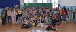 Grupa uczniów Szkoły Podstawowej nr 9 w Ełku pozuje wspólnie do zdjęcia w holu. Dzieci w różnym wieku stoją i siedzą razem,, niektóre trzymają książeczki i nagrody. Atmosfera jest radosna i żywa, co świadczy o udanym wydarzeniu edukacyjnym.