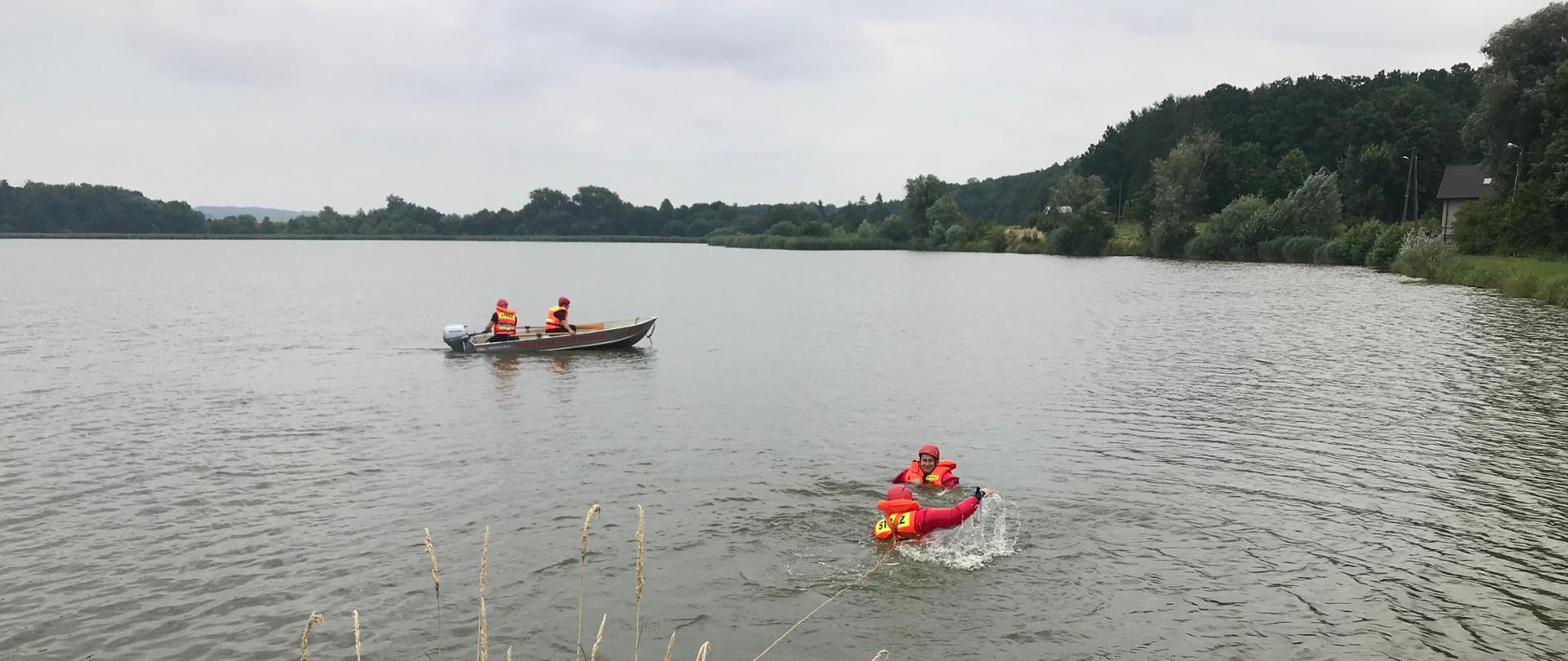 Strażak zabezpieczony w linkę asekuracyjną próbuje dotrzeć do osoby tonącej. W tle łódź ze strażakami pływająca po akwenie.
