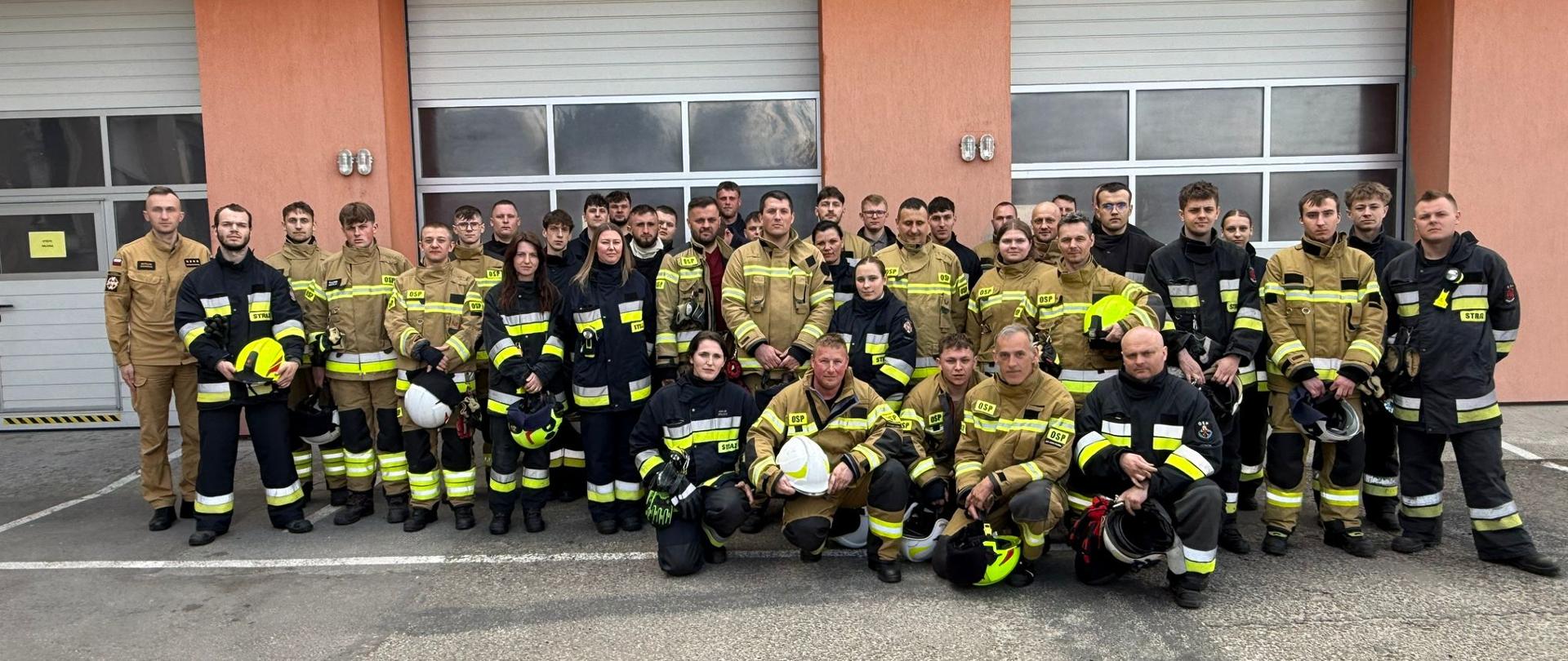 Strażacy ustawieni w kilku szeregach stoją ustawieni w na tle garażów. Ubrani są w ubrania specjalne koloru czarnego i pisakowego w rękach trzymają hełmy