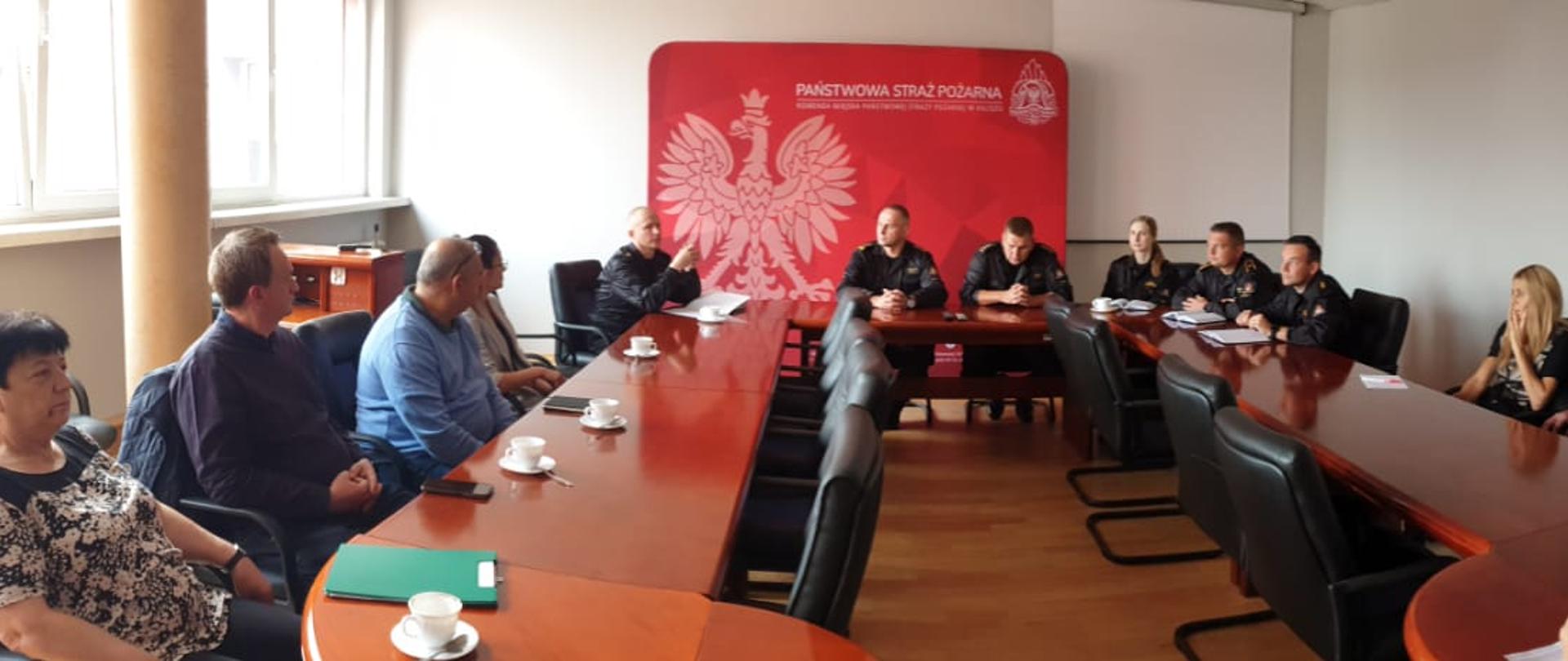 Na zdjęciu zamknięte pomieszczenie na terenie KM PSP w Kaliszu. W pomieszczeniu stoły i krzesła. Przy stołach ludzie. Zdjęcie w ciągu dnia. Kolor stołu brązowy. Wśród ludzi widać funkcjonariuszy KM PSP Kalisz.