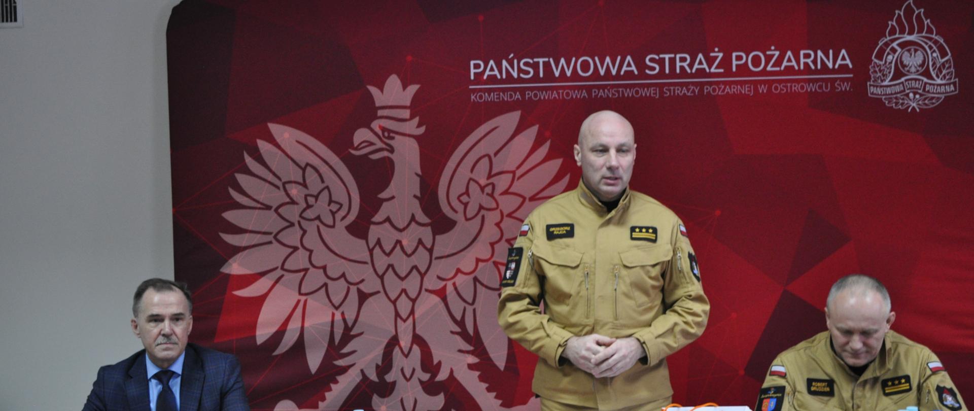 Na zdjęciu w sali konferencyjnej Komendy Powiatowej PSP w Ostrowcu Świętokrzyskim widać stojącego za stołem i zabierającego głos Zastępcę Świętokrzyskiego Komendanta Wojewódzkiego PSP w Kielcach st. bryg. Grzegorza Rajcę.