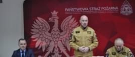 Na zdjęciu w sali konferencyjnej Komendy Powiatowej PSP w Ostrowcu Świętokrzyskim widać stojącego za stołem i zabierającego głos Zastępcę Świętokrzyskiego Komendanta Wojewódzkiego PSP w Kielcach st. bryg. Grzegorza Rajcę.