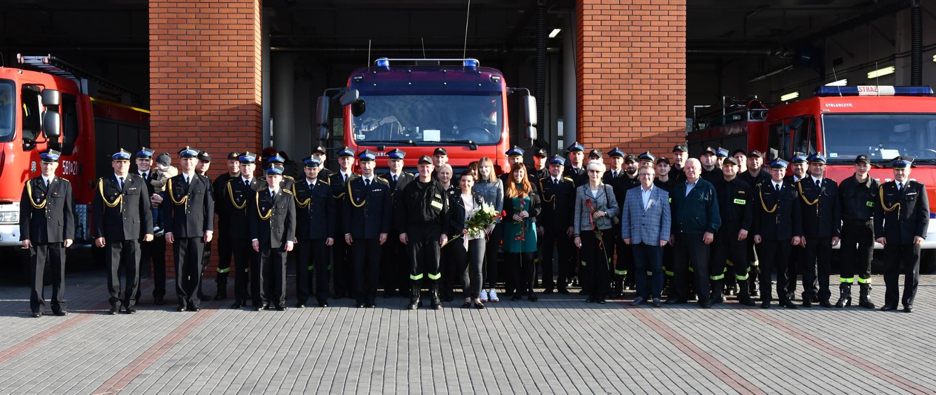 Na zdjęciu strażacy ubrani w mundury wyjściowe kolor granatowego oraz strażacy w ubraniach koszarowych stoją na tle samochodów pożarniczych za nimi budynek komendy