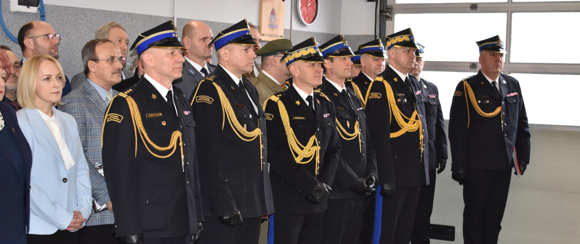 grupa ludzi stoi pod ścianą garażu, w pierwszym rzędzie stoją strażacy na baczność