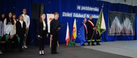 Zdjęcie przesdtawia grupę uczniów stojących na scenie. W tle wisi granatowy baner z napisem 14 października Dzień Edukacji Narodowej.