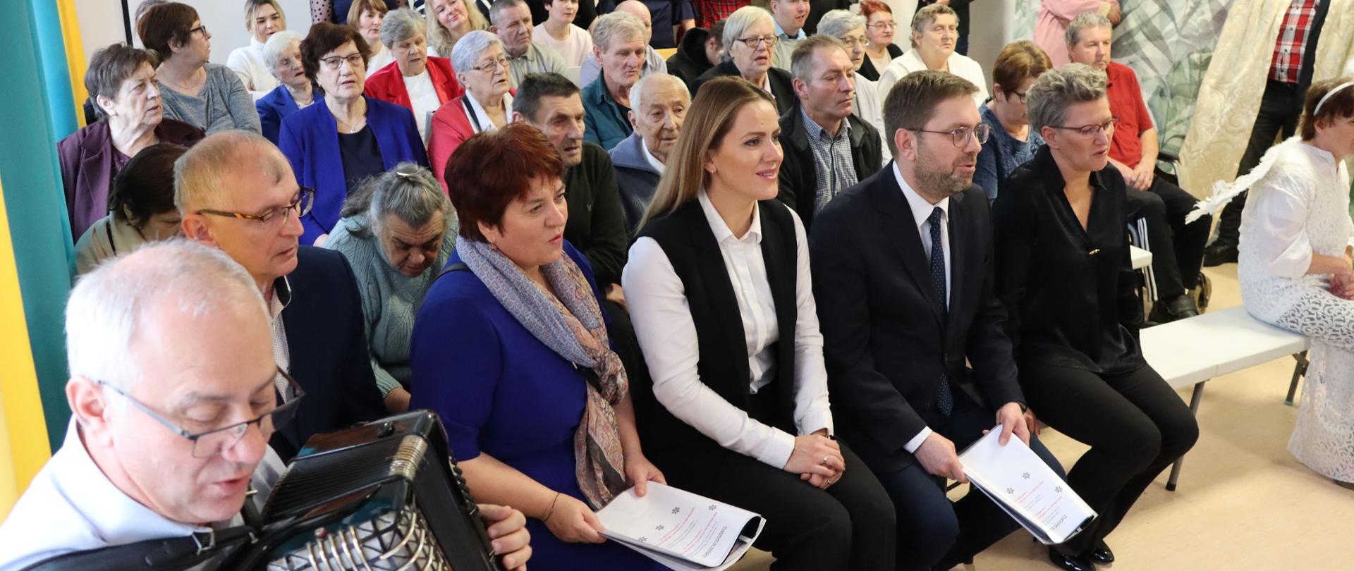 Kilkadziesiąt osób siedzi na sali obserwując przedstawienie. W dolnym lewym rogu pan gra na akordeonie.