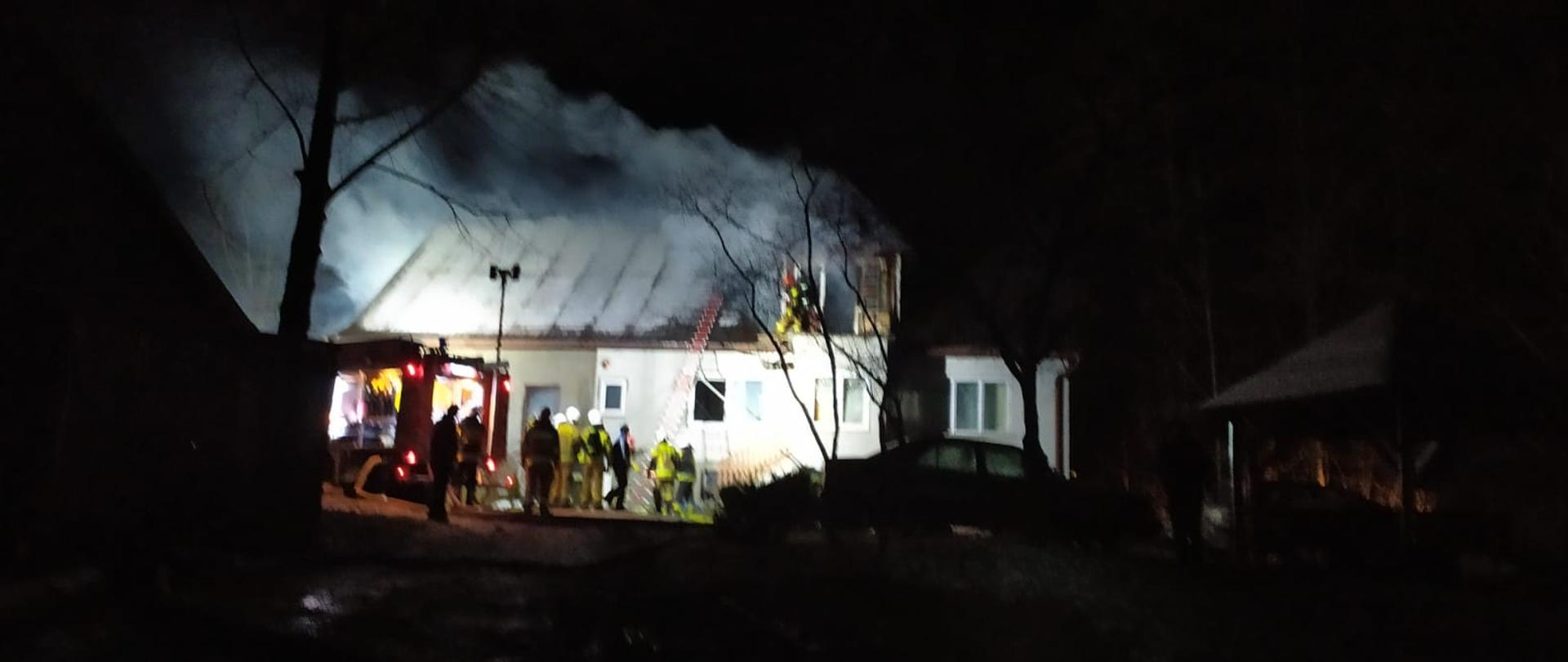 Zdjęcie wykonane w porze nocnej przedstawia budynek mieszkalny z nad którego unoszą się kłęby dymu. Przed obiektem stoi samochód strażacki, grupa ratowników prowadzących działania gaśnicze. O budynek oparta jest drabina i w górnej części również widzimy strażaków.