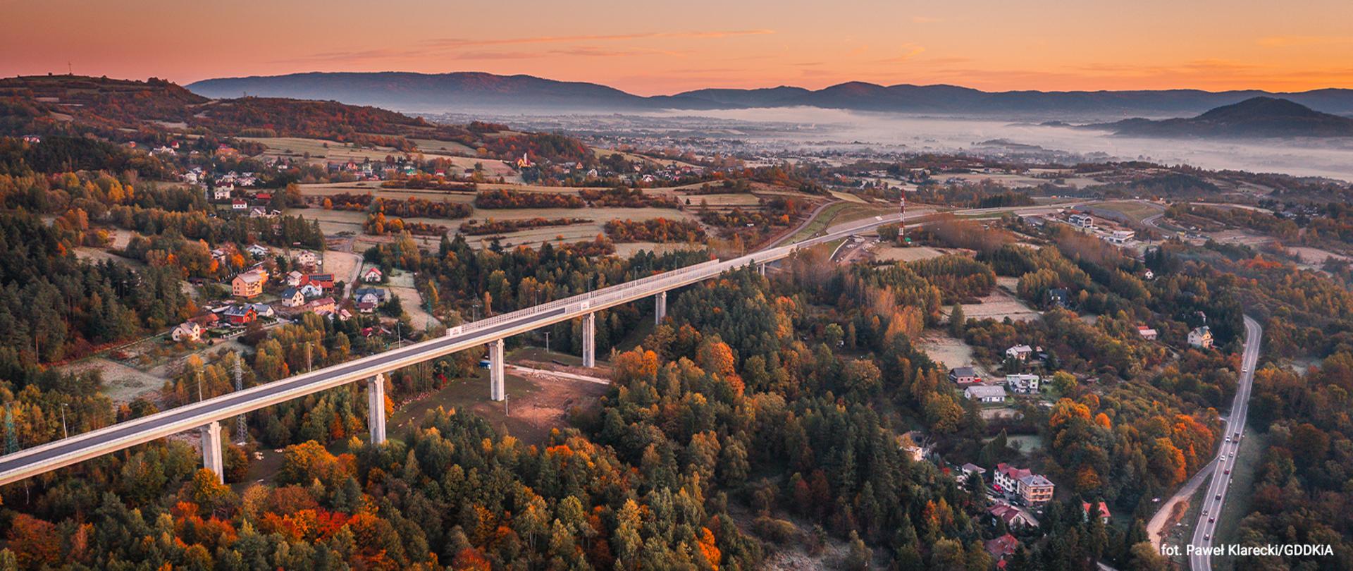 Widok na odcinek drogi ekspresowej S1 Przybędza–Milówka, przebiegający przez teren górski. Na zdjęciu widać trasę z estakadą lub węzłem drogowym, otoczoną zabudową, polami i lasami.