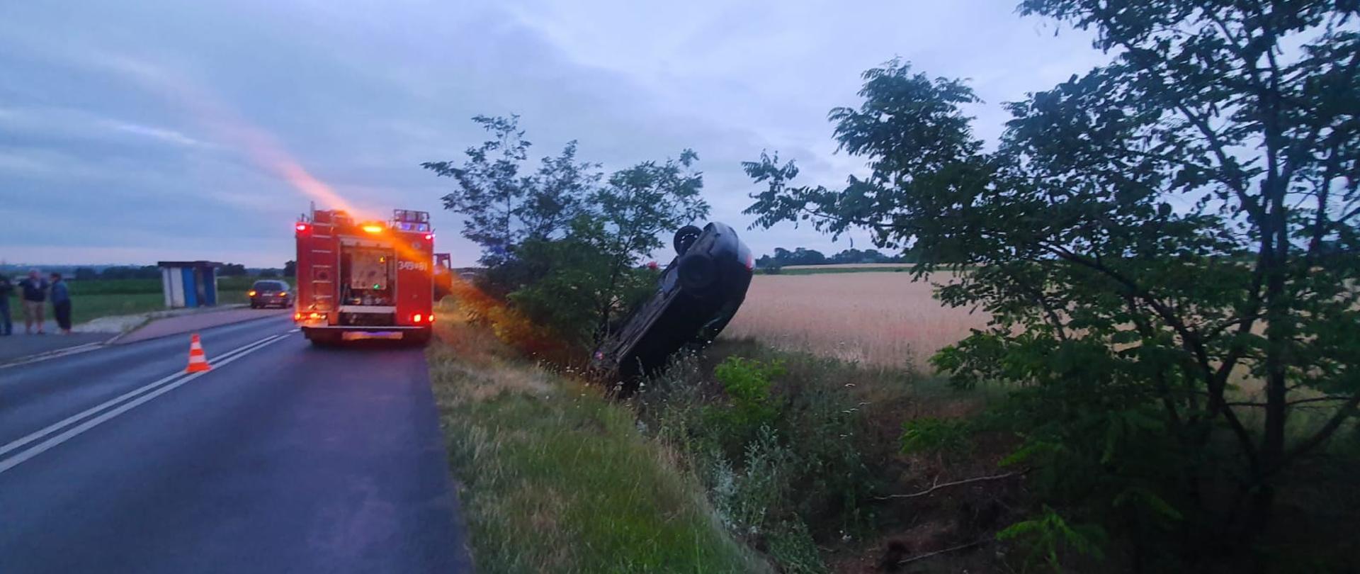 Na zdjęciu widać uszkodzony pojazd. Droga asfaltowa. Po bokach pola uprawne. Duże zachmurzenie. Zdjęcie w ciągu dnia. Na środku samochód ratowniczo-gaśniczy, czerwony.